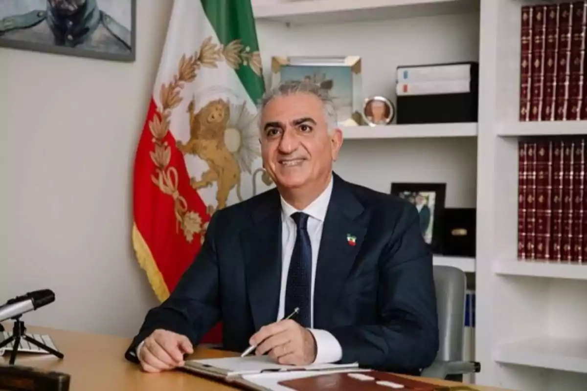 Hombre de cabello canoso con traje oscuro sentado en un escritorio, sosteniendo una pluma, con una bandera detrás y estanterías con libros a su alrededor
