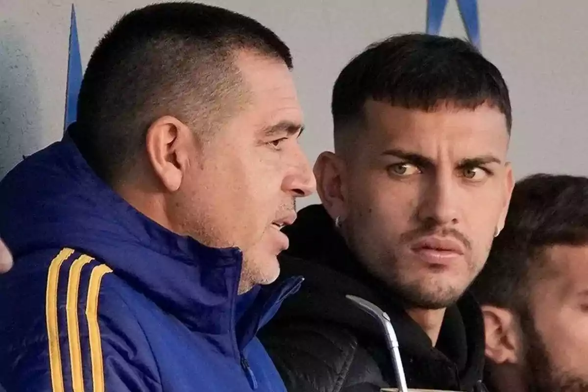 Two men are talking in the foreground while one of them holds a mate and the other wears a blue jacket with yellow stripes. Two men are talking in the foreground while one of them holds a mate and the other wears a blue jacket with yellow stripes.