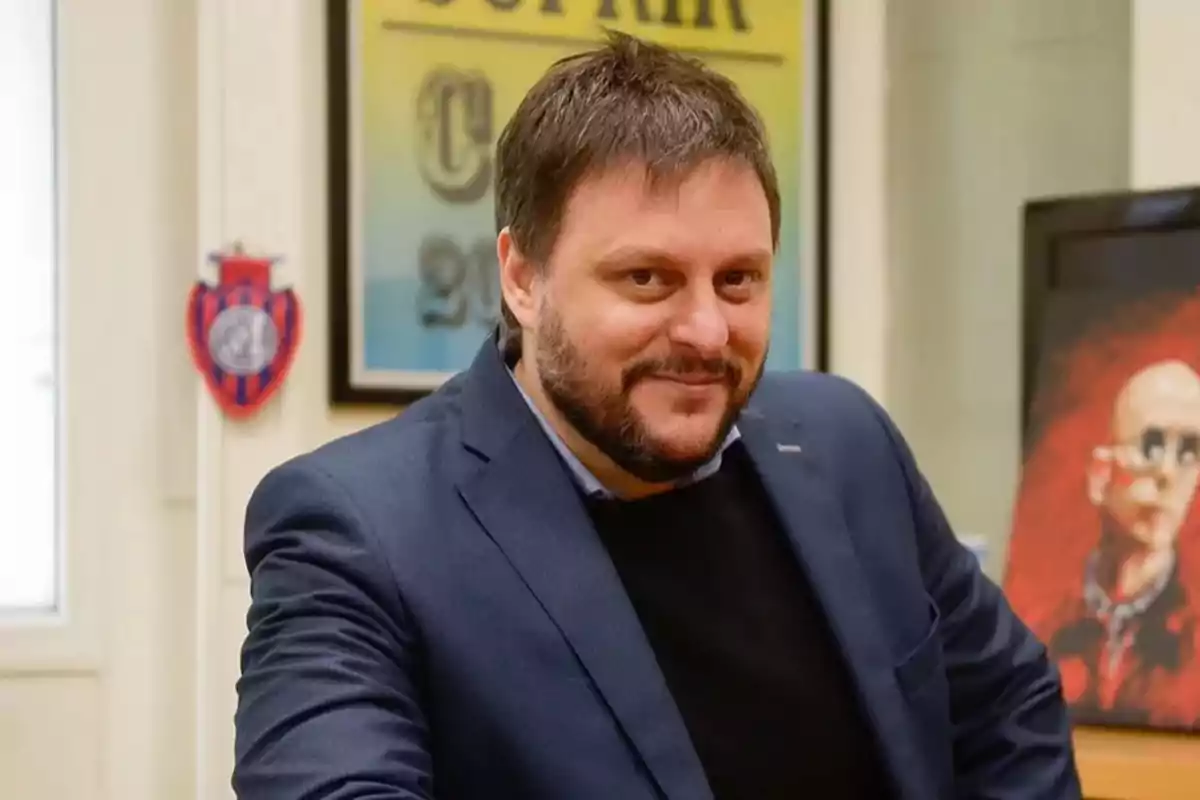 Un hombre con barba y cabello oscuro, vestido con un traje azul, sonríe mientras está en una oficina con decoraciones en la pared.