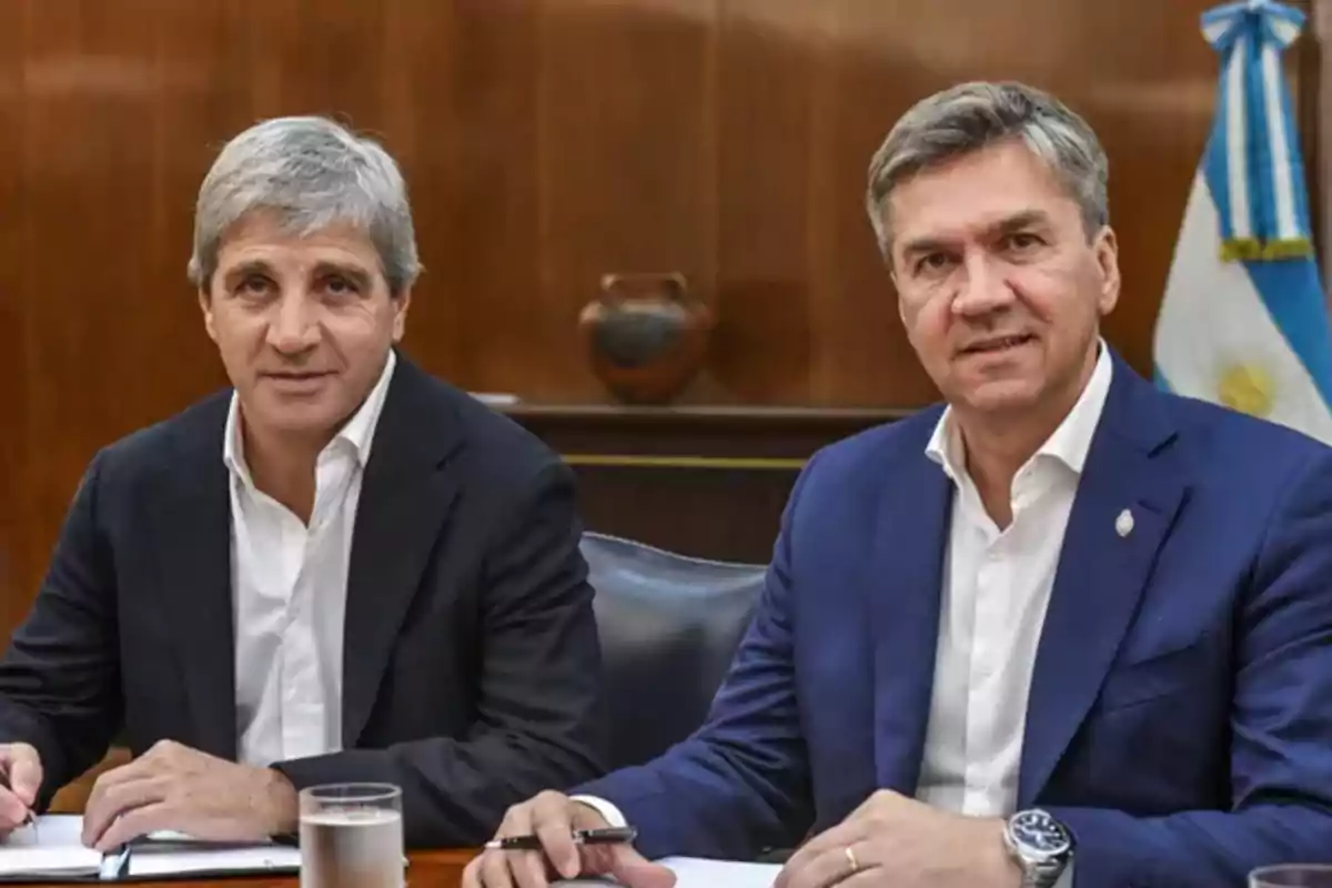 Dos hombres en trajes formales sentados en una mesa con documentos y un vaso de agua, con una bandera argentina en el fondo.