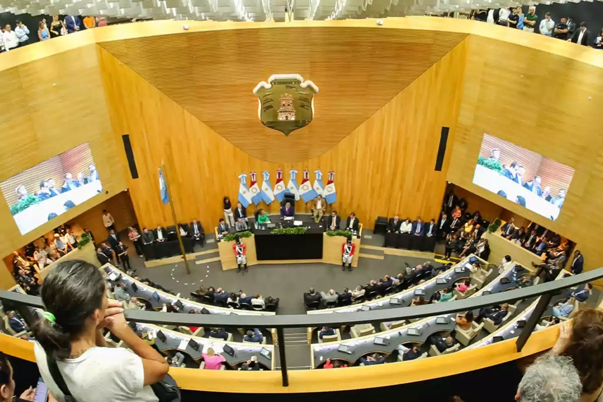 Vista panorámica del interior de un recinto legislativo con personas sentadas en bancas semicirculares, autoridades en el estrado principal, banderas argentinas y pantallas mostrando la sesión