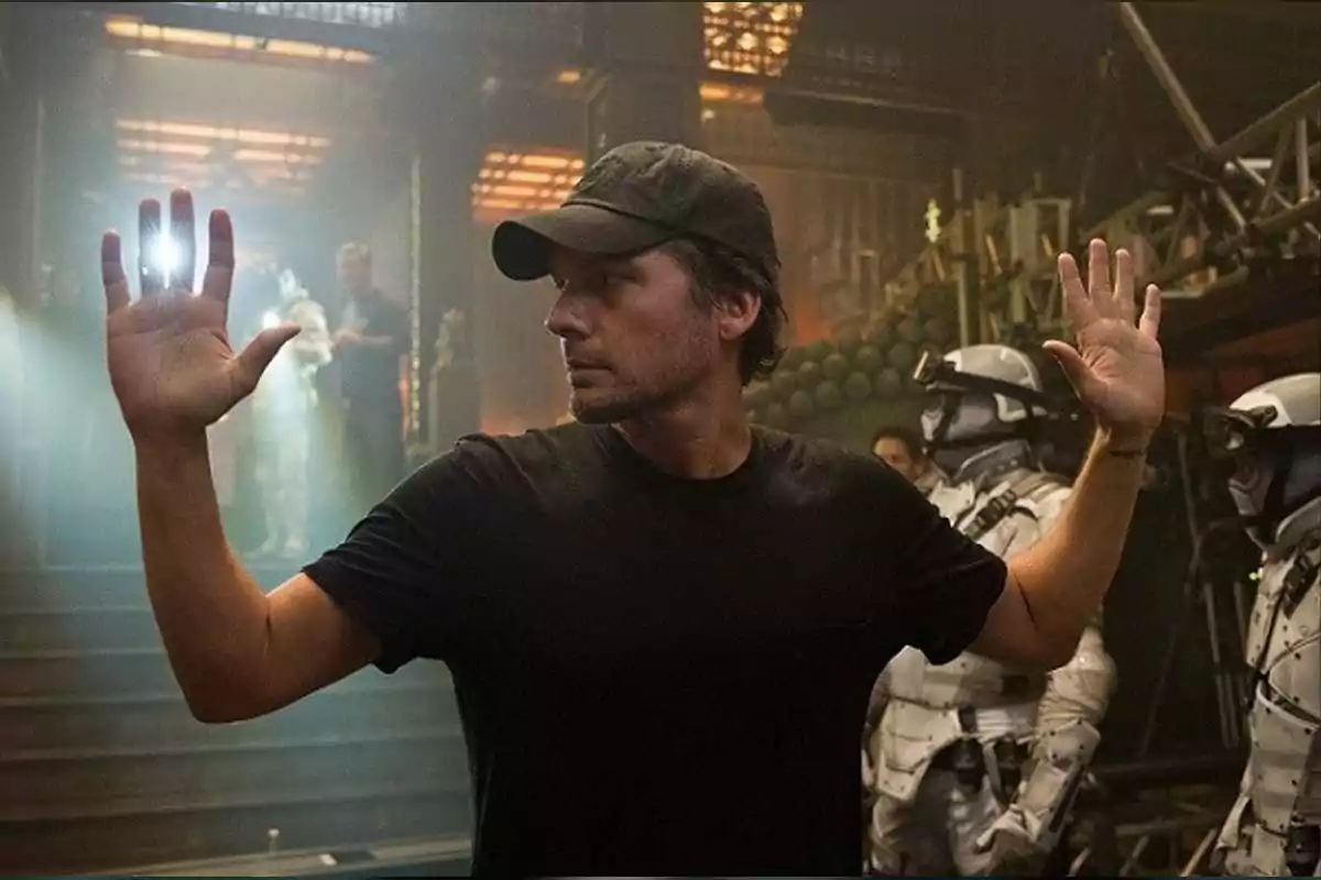 A man in a cap and black t-jersey raises his hands on a film set, while a flash of light emanates from his left hand and two people in futuristic suits are around him.