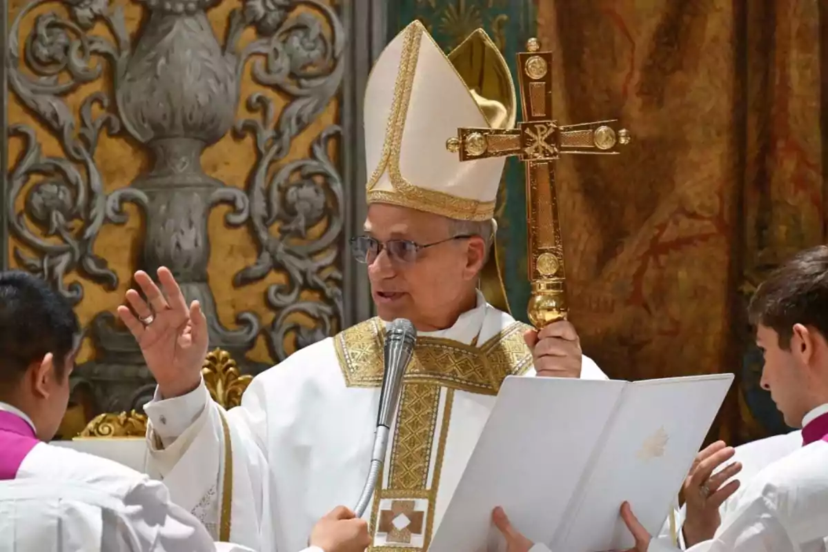 Un clérigo católico con vestimenta ceremonial sostiene un crucifijo dorado mientras realiza una bendición.