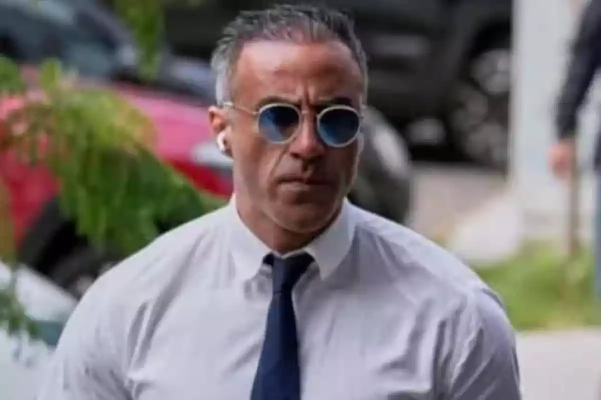 A man with sunglasses and headphones walks outdoors wearing a white jersey and a blue tie.