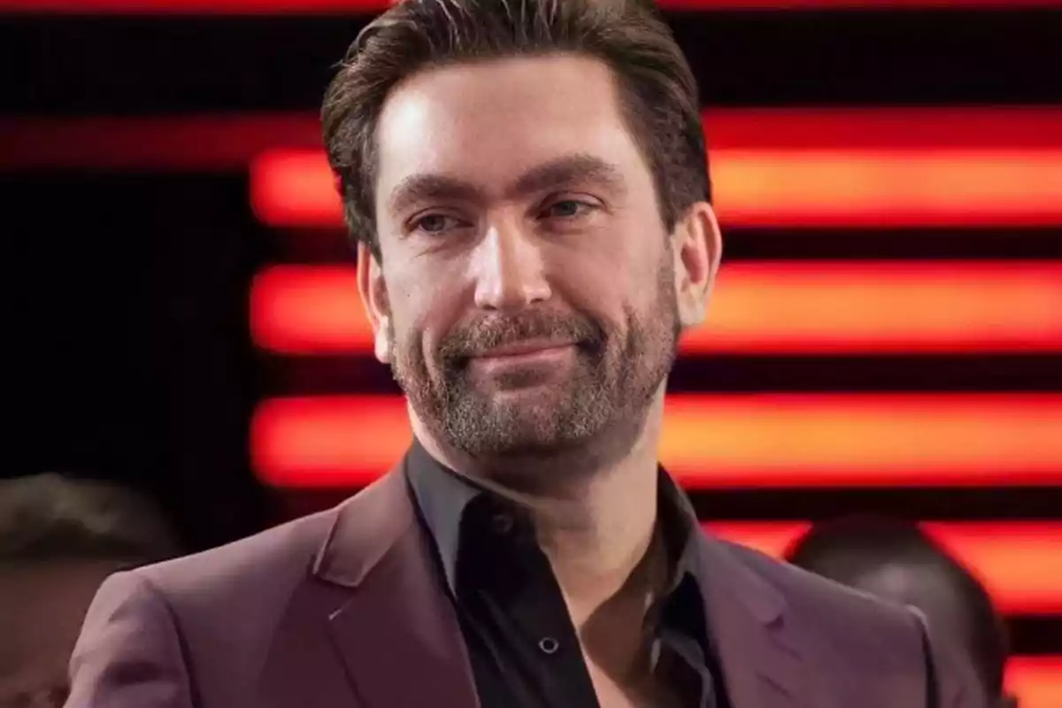 Hombre de cabello oscuro y barba corta con saco marrón y camisa negra, fondo con luces rojas horizontales. Hombre de cabello oscuro y barba corta con saco marrón y camisa negra, fondo con luces rojas horizontales.
