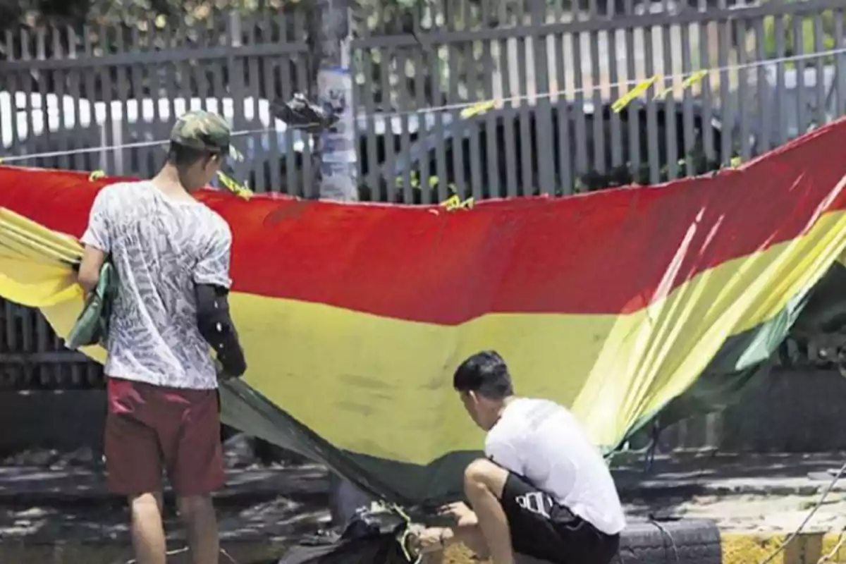 Dos personas ajustan una gran tela con los colores rojo, amarillo y verde en un espacio al aire libre.