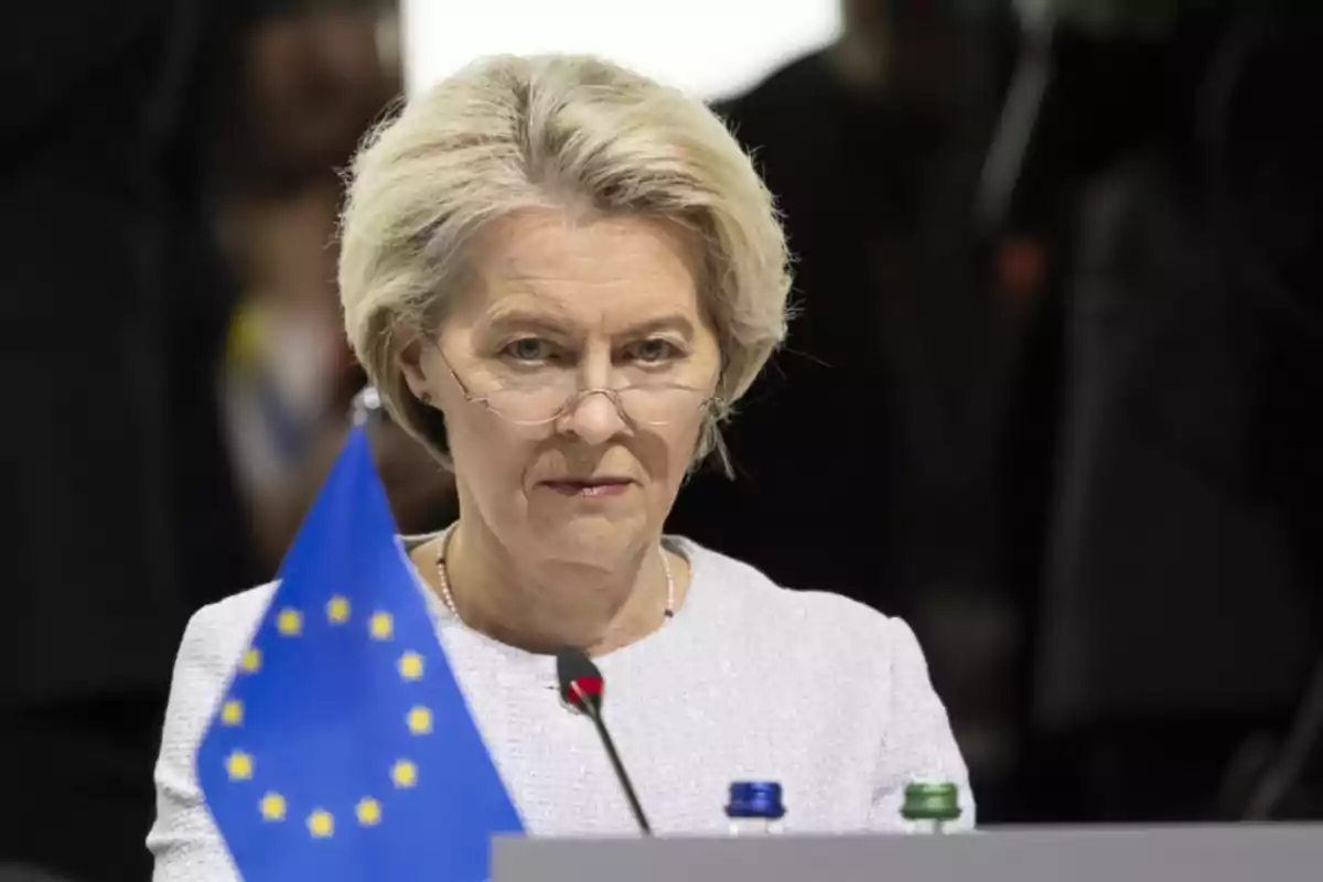 A person with glasses and blonde hair is sitting in front of a microphone, with a European Union flag in the foreground.