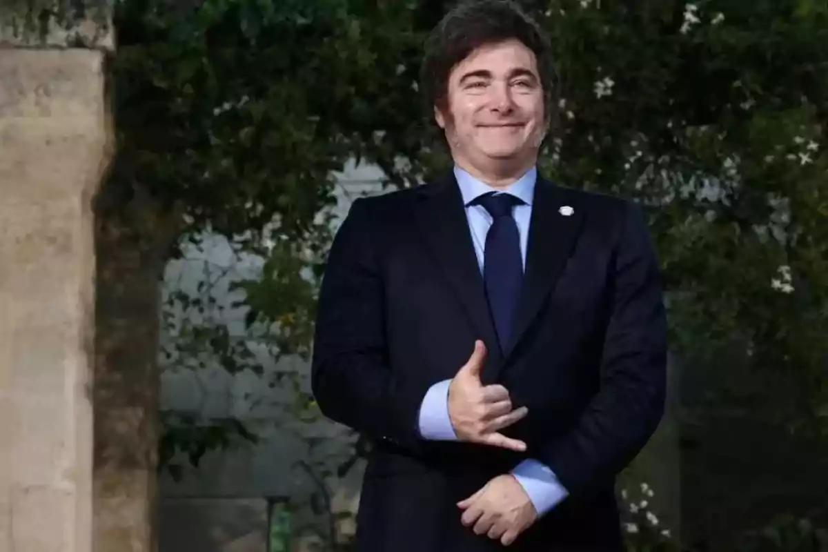 Un hombre con traje oscuro y corbata azul sonríe mientras hace un gesto con la mano frente a un fondo de vegetación.