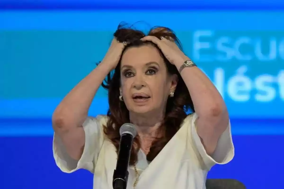 Mujer con expresión de sorpresa o preocupación, con las manos en la cabeza, frente a un micrófono y un fondo azul.