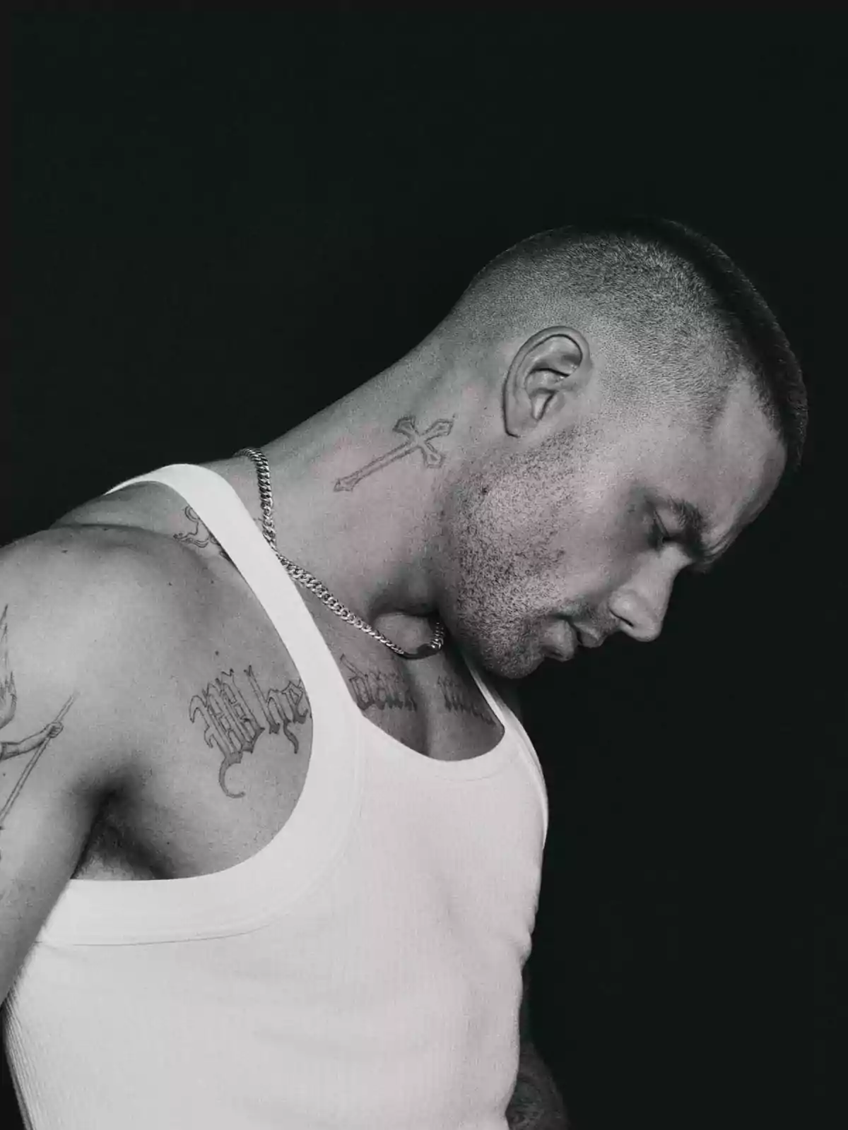Un hombre con el cabello corto y tatuajes visibles en el cuello y el brazo, vistiendo una camiseta sin mangas blanca, en un fondo oscuro. Un hombre con el cabello corto y tatuajes visibles en el cuello y el brazo, vistiendo una camiseta sin mangas blanca, en un fondo oscuro.