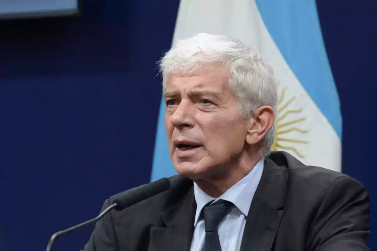 Un hombre de cabello canoso hablando en un micrófono con una bandera argentina de fondo.