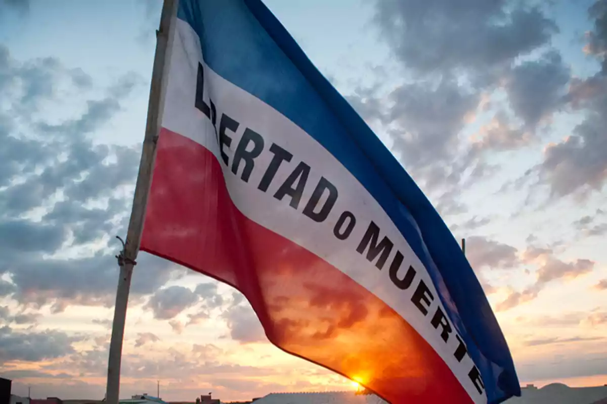 Bandera ondeando con la frase "Libertad o Muerte" al atardecer.