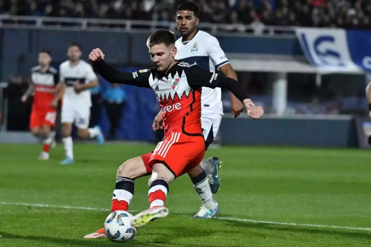 Un jugador de fútbol con uniforme rojo y negro está a punto de patear el balón mientras es perseguido por un jugador del equipo contrario vestido de blanco durante un partido.