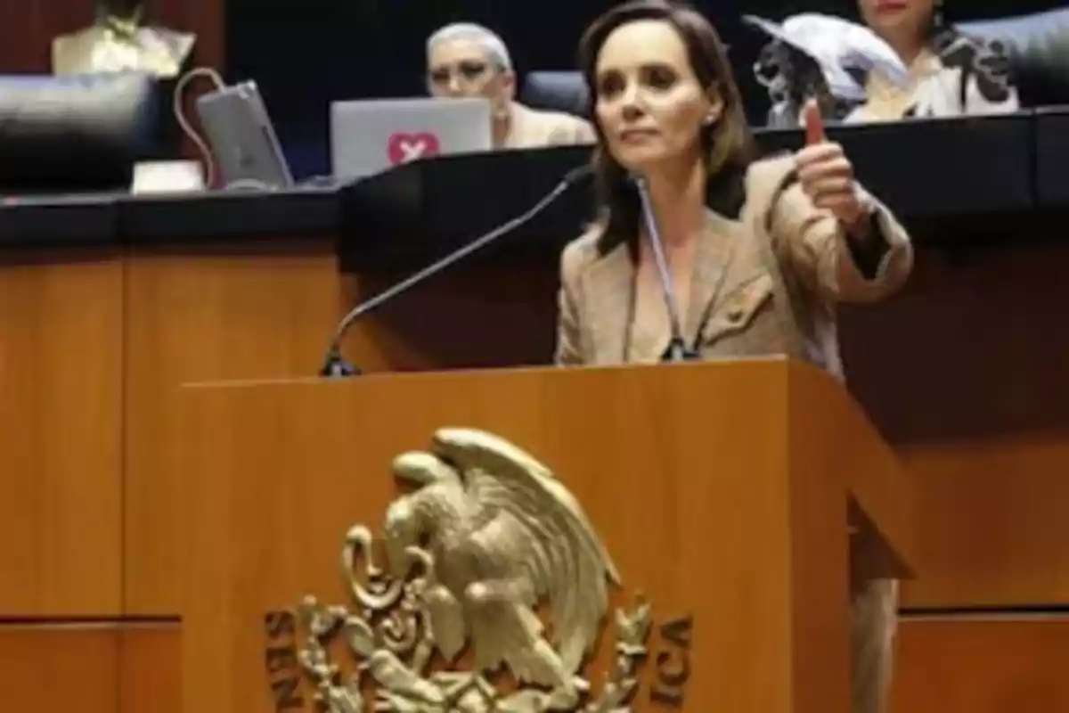 Una persona en un podio de madera con el escudo nacional de México, levantando el pulgar mientras habla en un micrófono, con otras personas y computadoras portátiles al fondo.