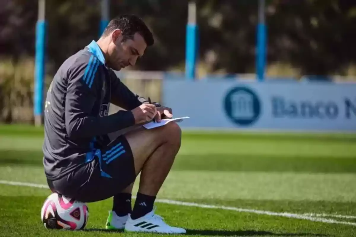 A man in sportswear is crouched over a soccer ball taking notes in a notebook on a training field.