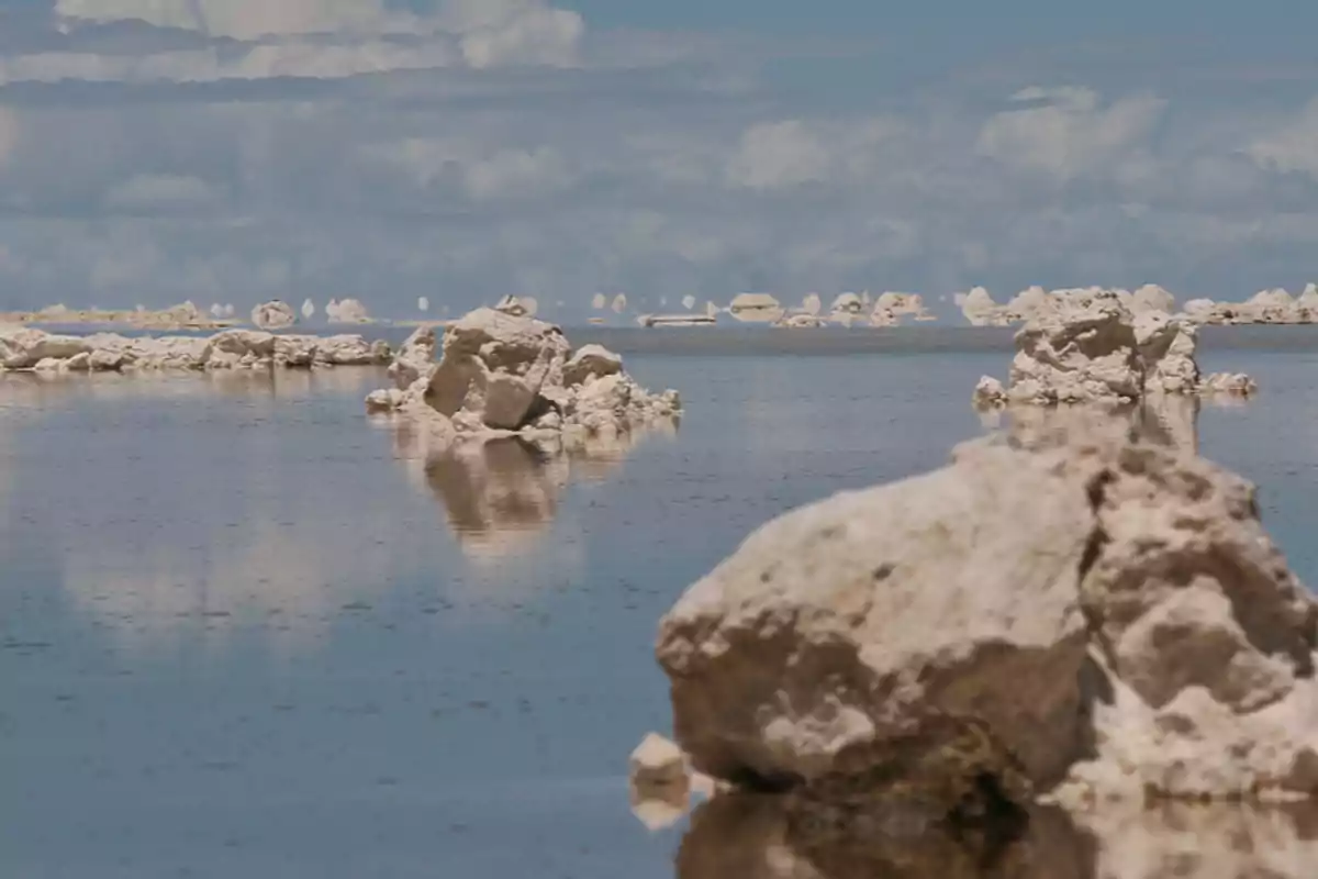 Una vista de un paisaje desértico con formaciones rocosas blancas reflejadas en una superficie de agua tranquila bajo un cielo parcialmente nublado.