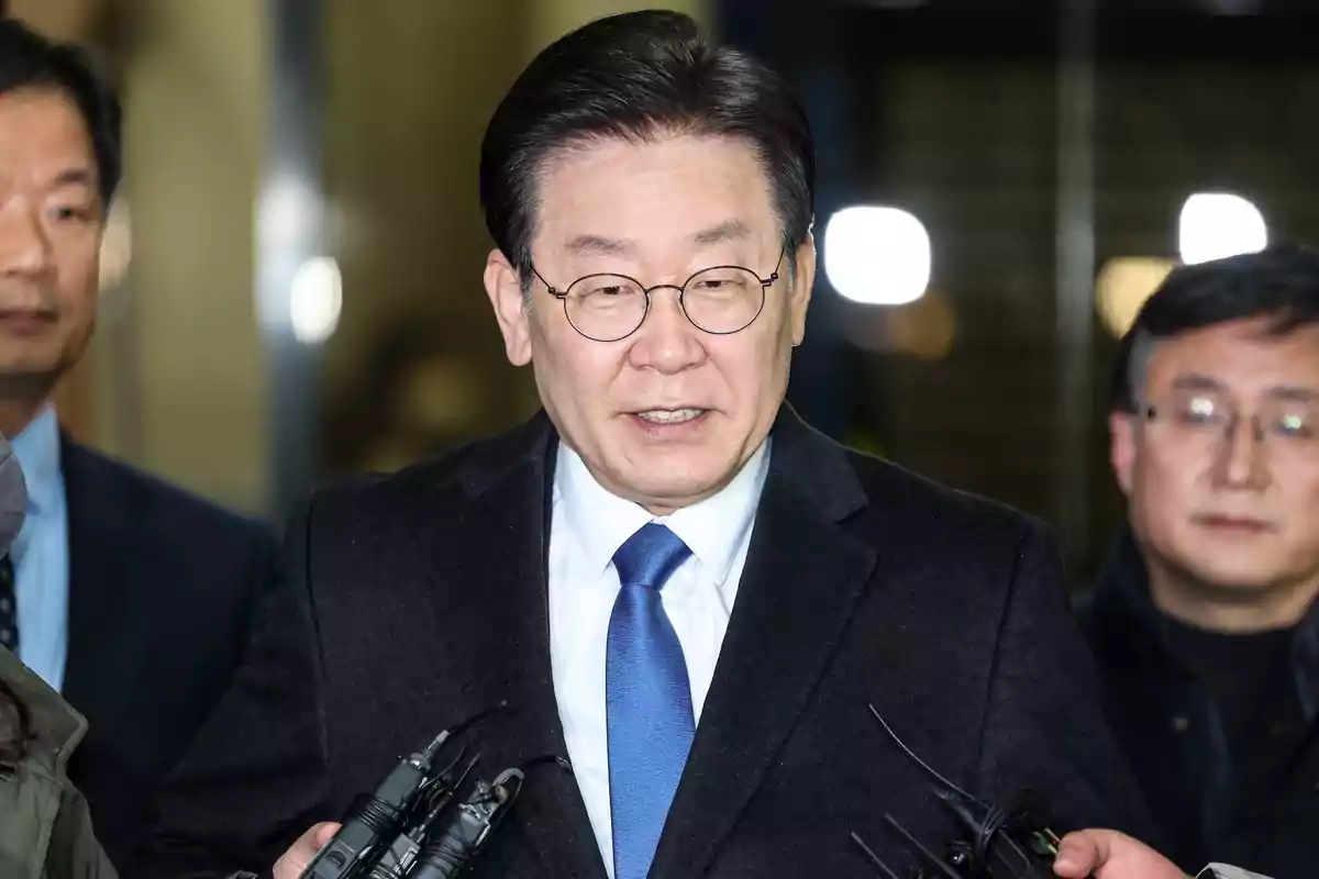 Dark-haired man with round glasses wearing a blue suit and tie surrounded by people and microphones in a well-lit indoor setting