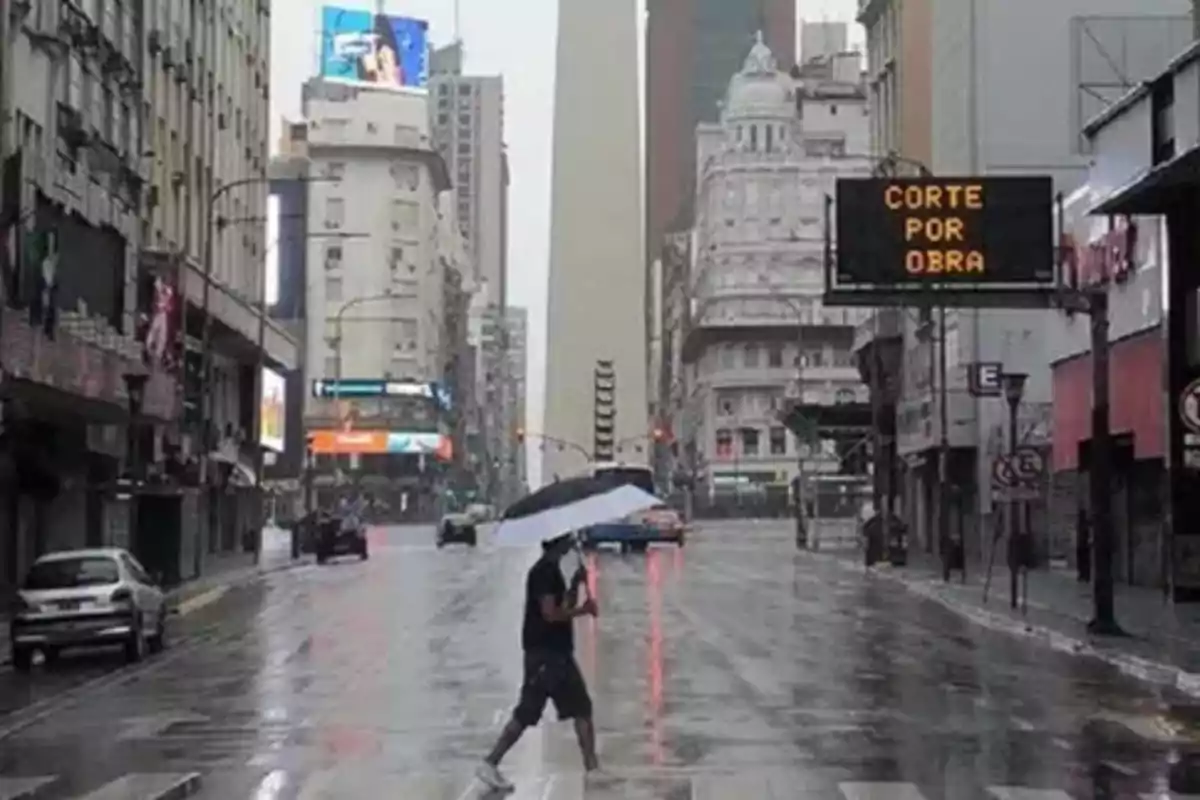 Persona cruzando una calle mojada con un paraguas en un día lluvioso en una ciudad con edificios altos y un cartel de 