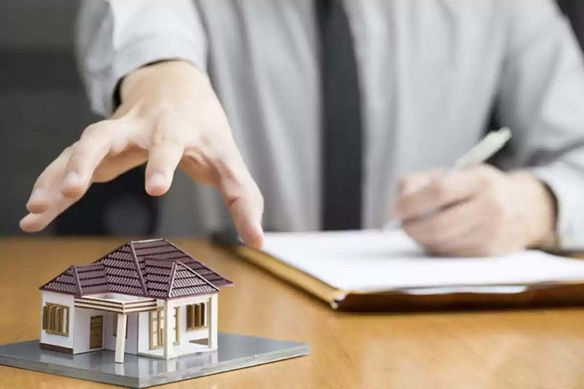 Una mano extendida sobre una maqueta de casa mientras una persona escribe en un documento.