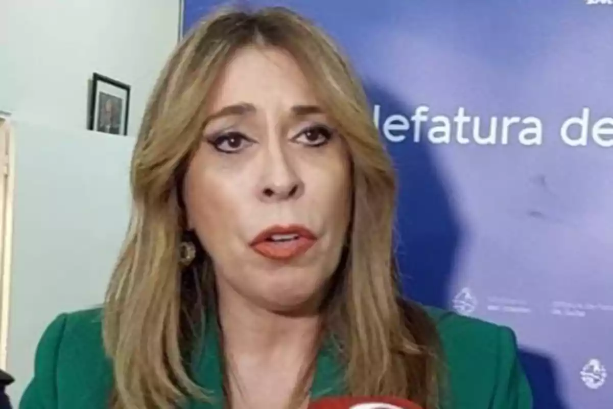Mujer de cabello rubio y largo hablando frente a un micrófono con fondo azul de oficina institucional Mujer de cabello rubio y largo hablando frente a un micrófono con fondo azul de oficina institucional