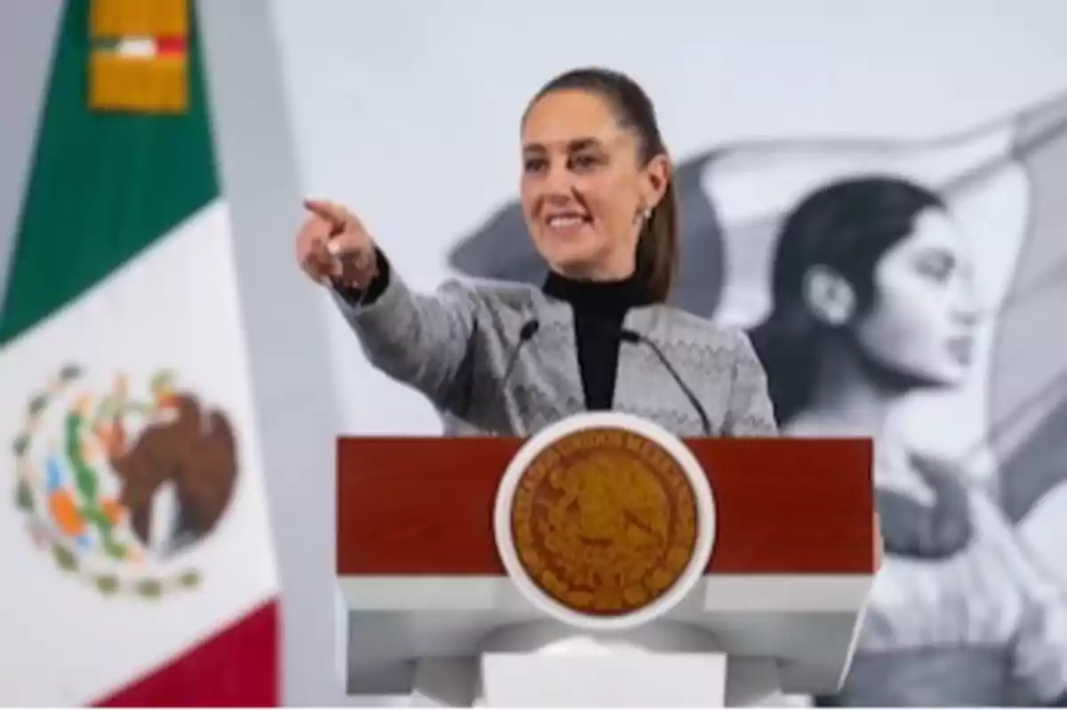 Una persona hablando en un podio con el escudo de México y una bandera mexicana al fondo.