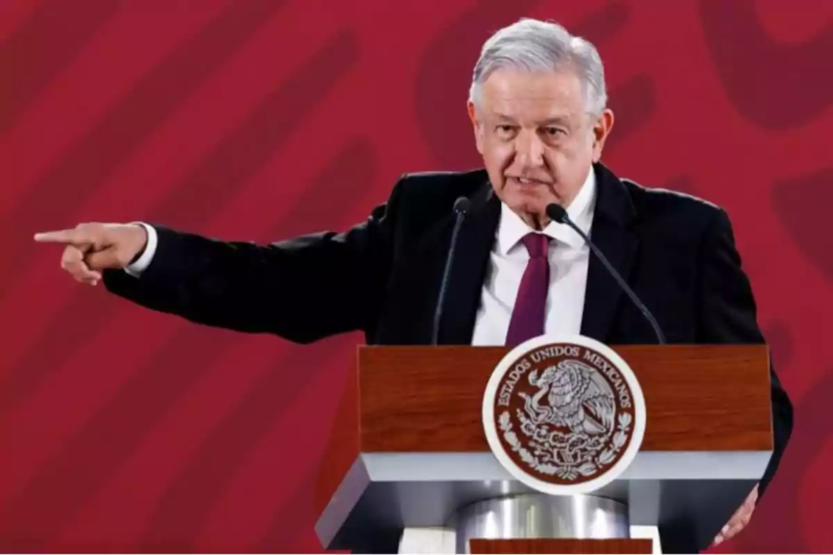 Un hombre de traje oscuro y corbata roja habla en un podio con el escudo de México, señalando hacia un lado.