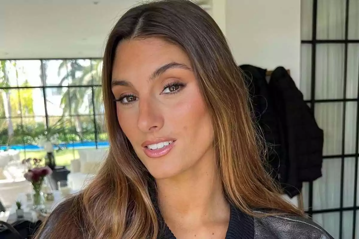 A person with long brown hair smiling indoors with large windows and greenery in the background. A person with long brown hair smiling indoors with large windows and greenery in the background.