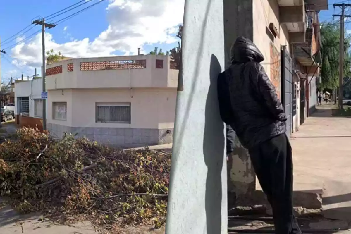 A hooded person leans against a wall on an urban street, while on the other side there is a corner house with fallen branches on the sidewalk.