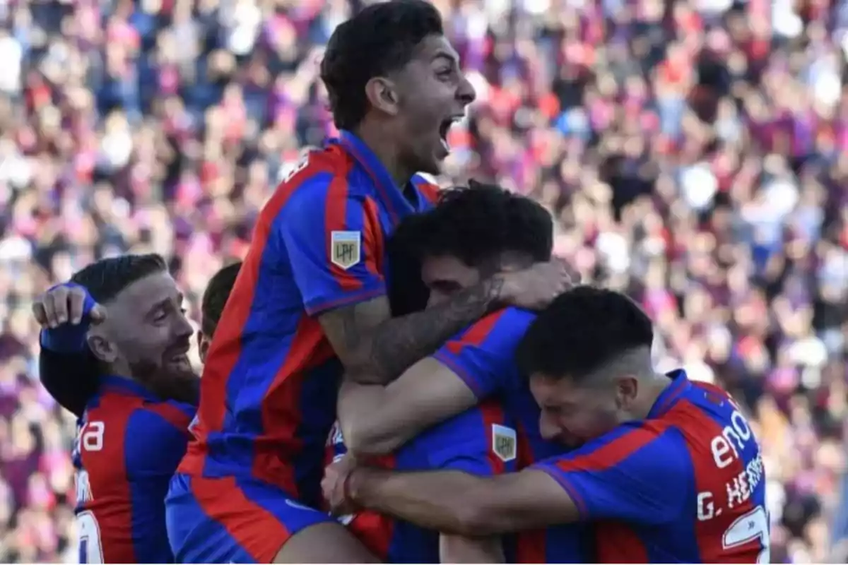 Jugadores de fútbol celebran un gol abrazándose en el campo, vistiendo uniformes azules y rojos, con una multitud de fondo. Jugadores de fútbol celebran un gol abrazándose en el campo, vistiendo uniformes azules y rojos, con una multitud de fondo.