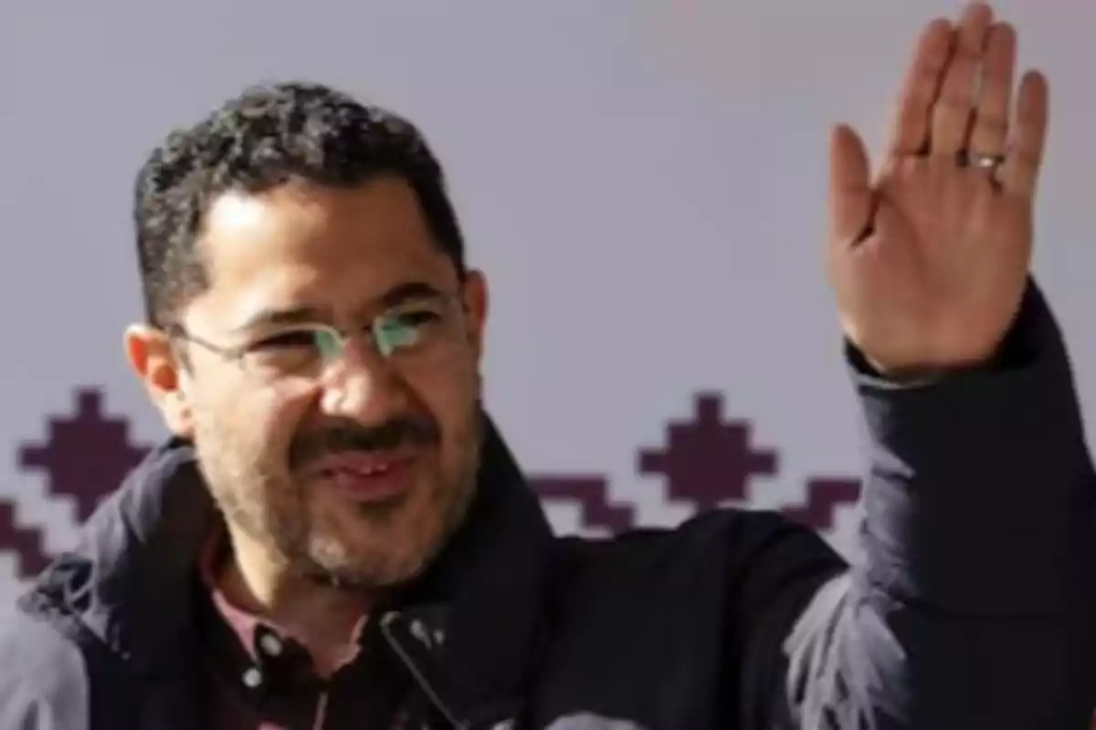 Un hombre con gafas y barba levanta la mano en señal de saludo, vistiendo una chaqueta oscura y con un fondo de diseño geométrico.