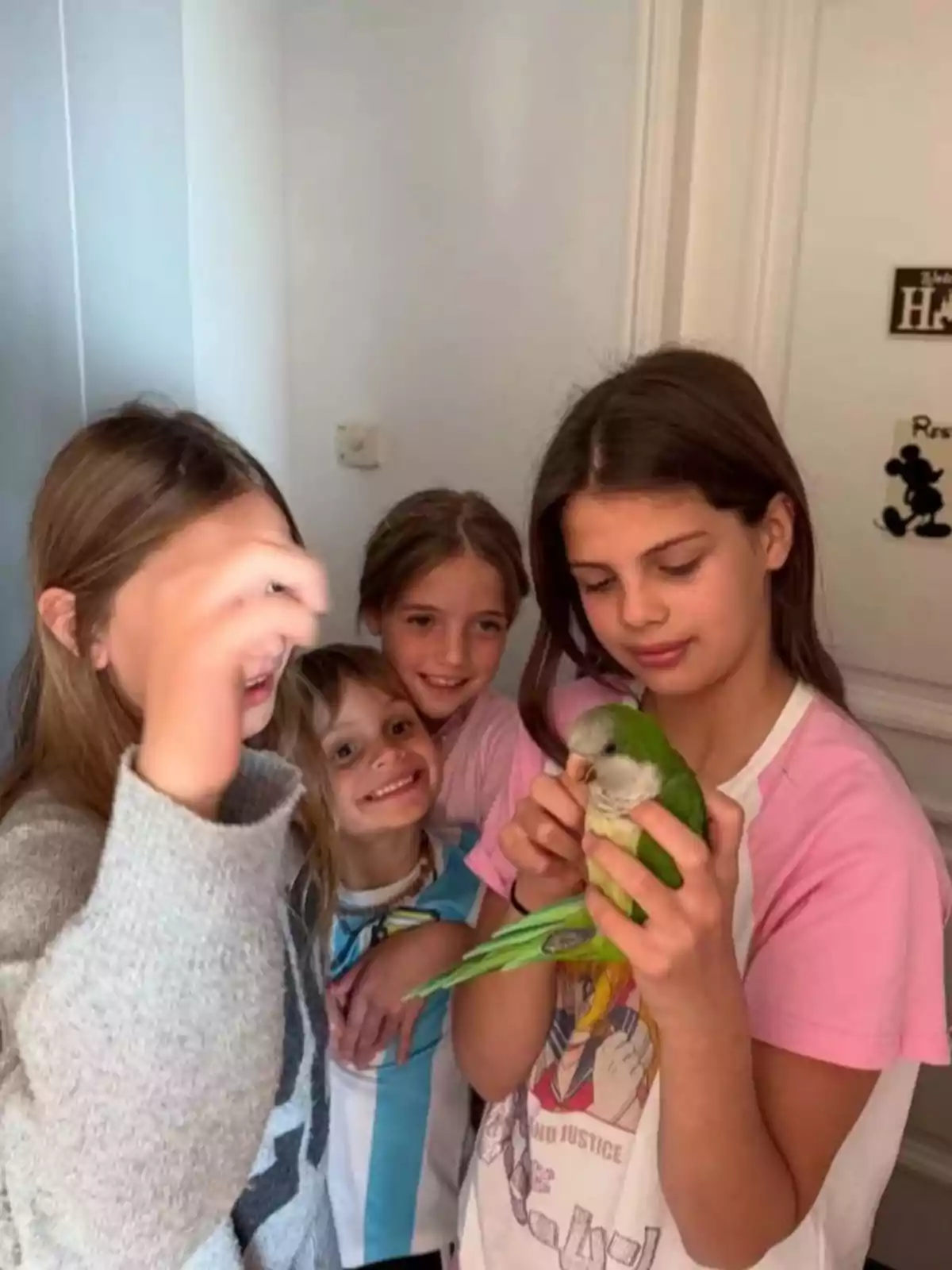 Four girls smile while one of them holds a green parrot in her hands inside a house.