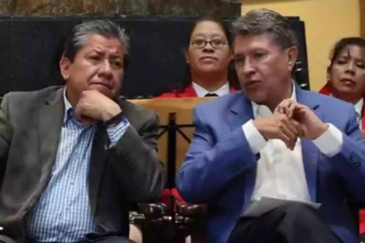 Dos hombres sentados conversando en un evento formal con personas en el fondo.