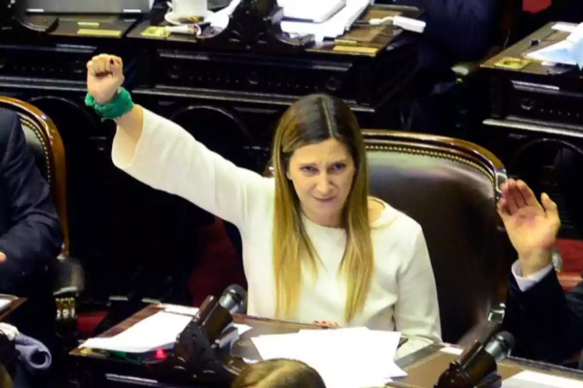 Una persona con el brazo levantado y un pañuelo verde en la muñeca, sentada en un entorno legislativo con papeles sobre la mesa. Una persona con el brazo levantado y un pañuelo verde en la muñeca, sentada en un entorno legislativo con papeles sobre la mesa.
