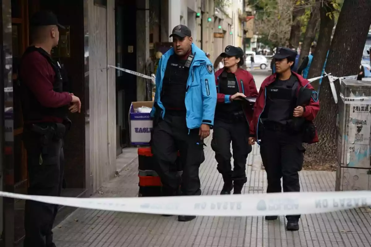 Un grupo de policías camina por una acera delimitada con cinta de seguridad en una zona urbana.