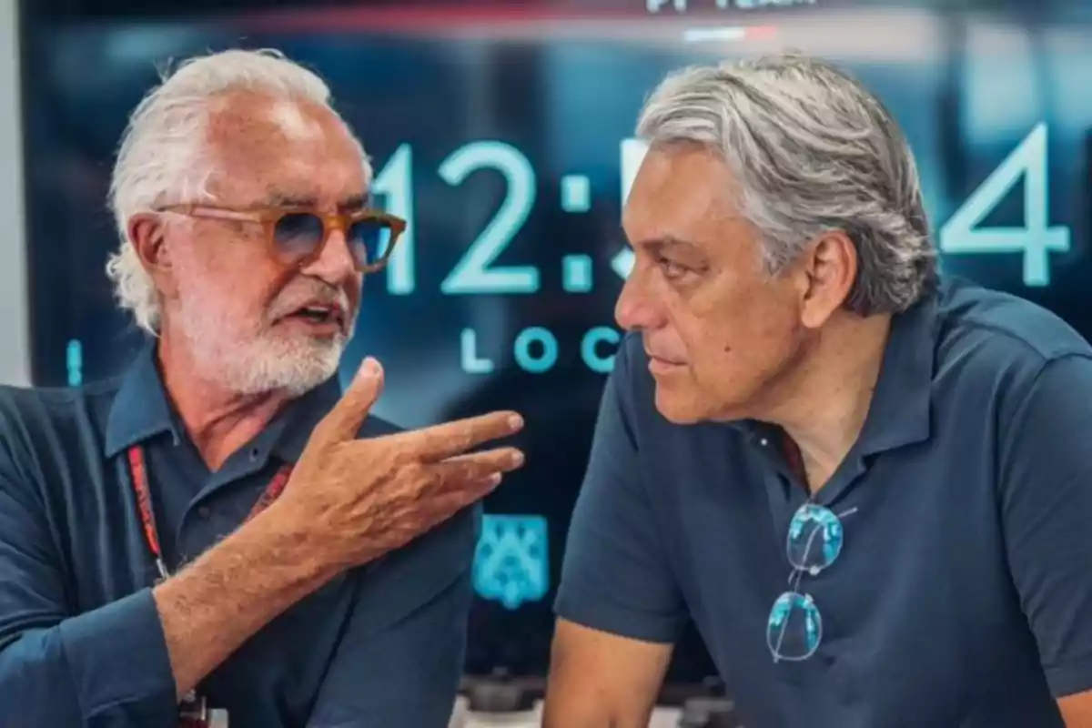 Two gray-haired men sit talking in front of a digital screen displaying the time. Two gray-haired men sit talking in front of a digital screen displaying the time.