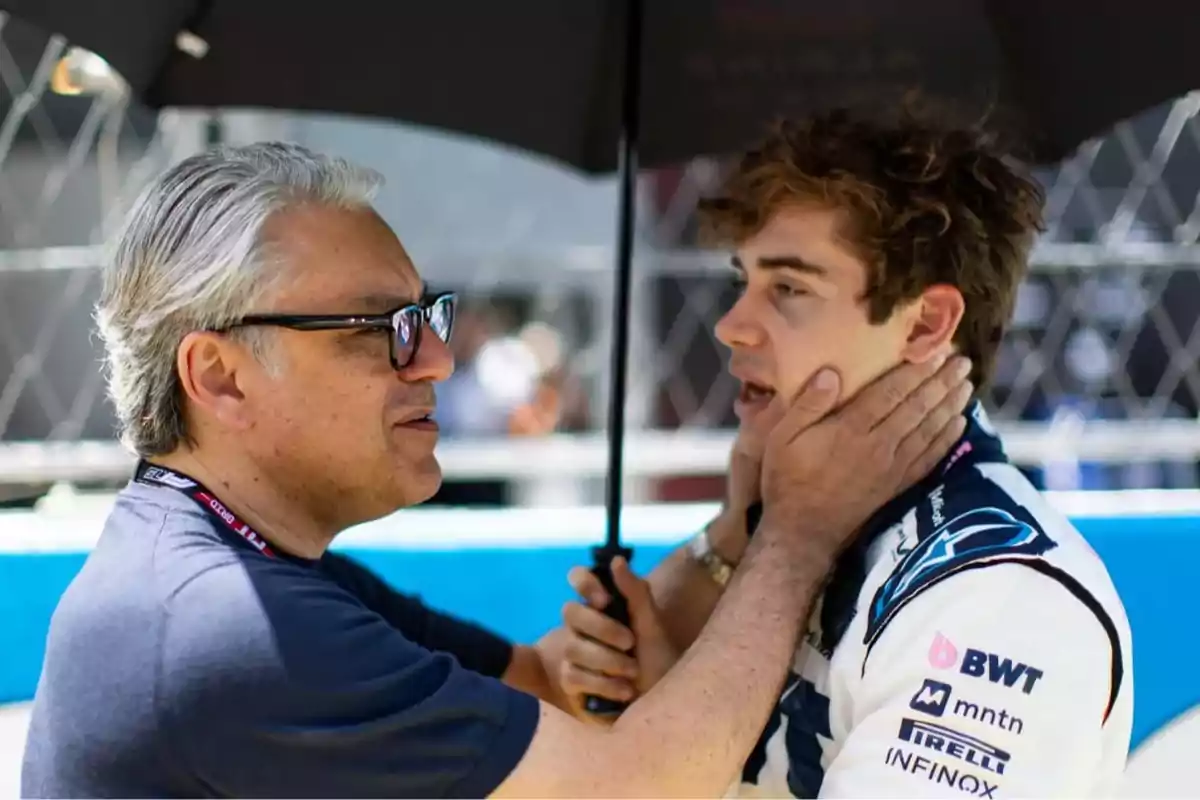 An older man holds the face of a young race car driver while both are under an umbrella in a motorsport competition setting. An older man holds the face of a young race car driver while both are under an umbrella in a motorsport competition setting.