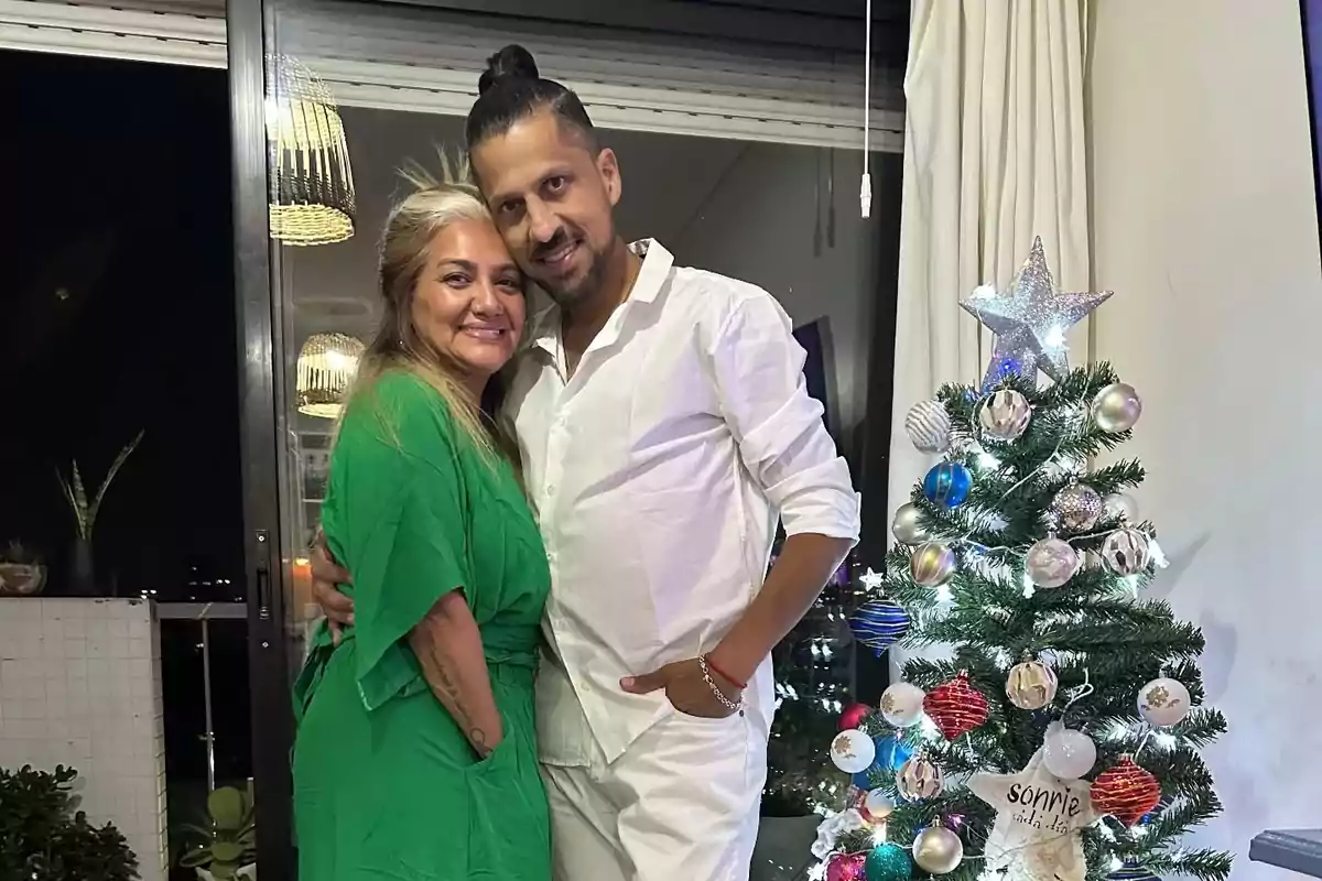 A smiling couple poses next to a Christmas tree decorated with lights and colorful ornaments in a well-lit room. A smiling couple poses next to a Christmas tree decorated with lights and colorful ornaments in a well-lit room.