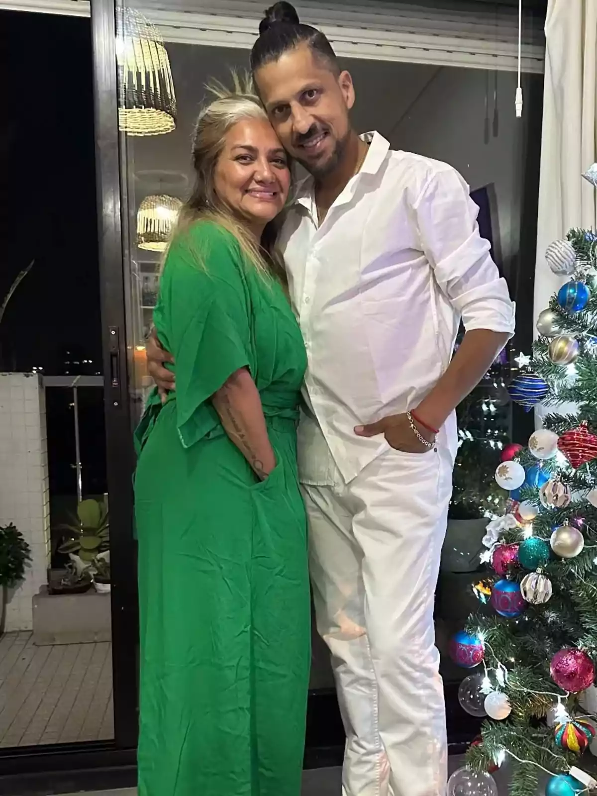 Una pareja sonriente posa junto a un árbol de Navidad decorado, ella lleva un vestido verde y él una camisa blanca, en un ambiente interior iluminado. Una pareja sonriente posa junto a un árbol de Navidad decorado, ella lleva un vestido verde y él una camisa blanca, en un ambiente interior iluminado.
