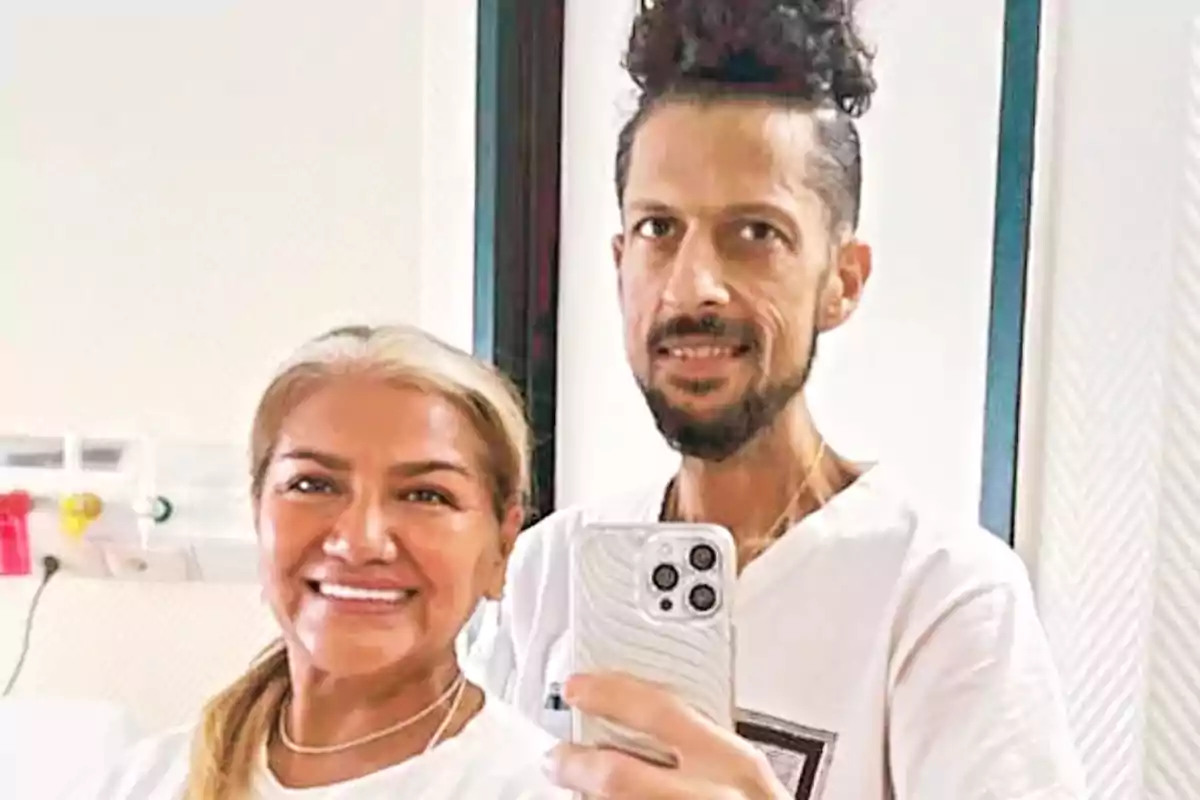 Two people smile while taking a selfie in front of a mirror; both are wearing white clothes and are in a well-lit indoor setting.