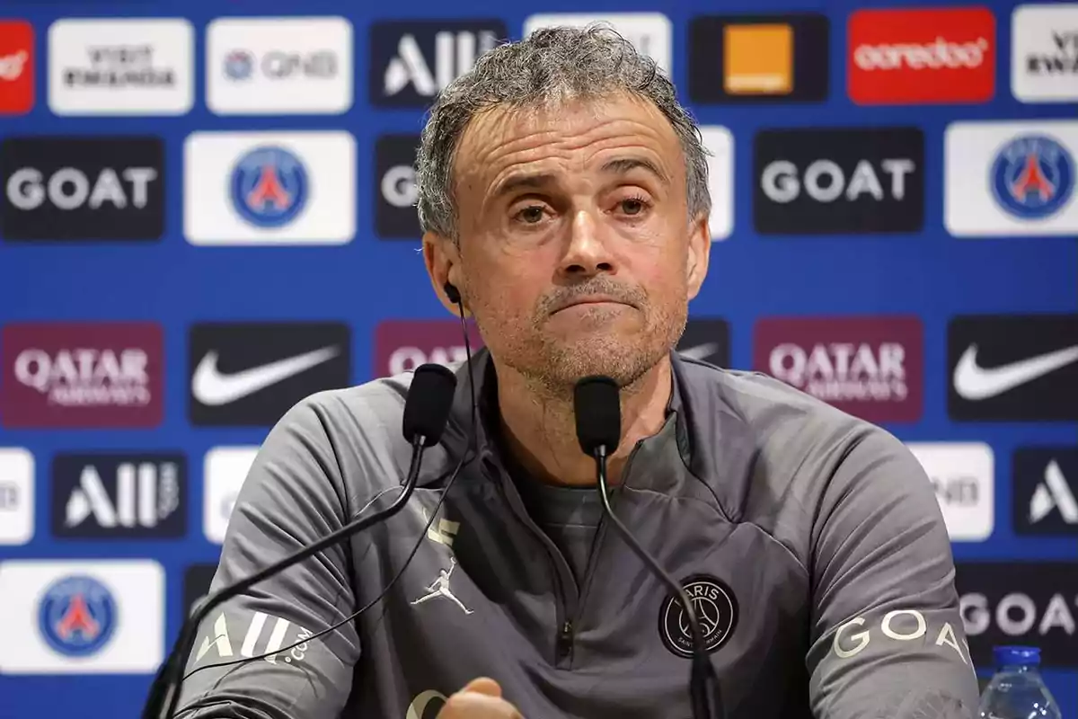 Entrenador del PSG en conferencia de prensa con micrófonos al frente y fondo de patrocinadores del club Entrenador del PSG en conferencia de prensa con micrófonos al frente y fondo de patrocinadores del club