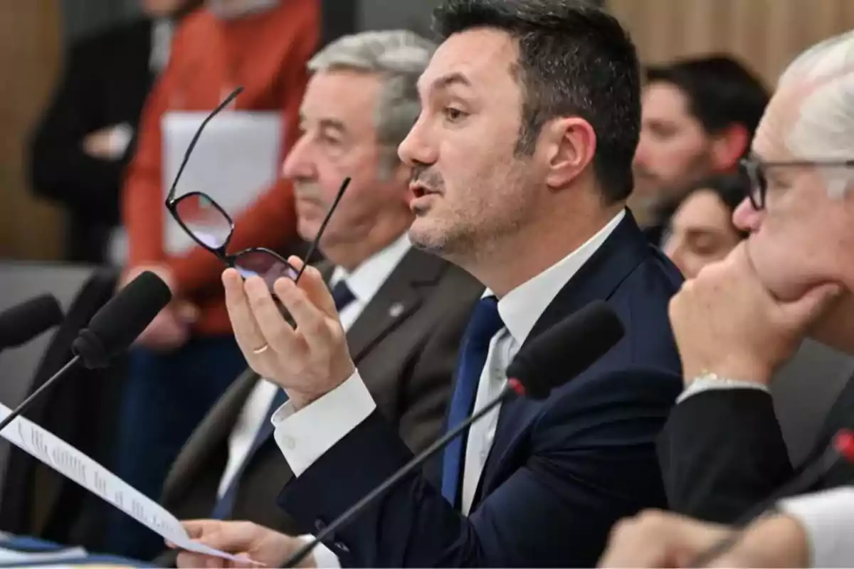 Man in a suit speaking at a meeting while holding glasses in his hand, surrounded by other people and microphones. Man in a suit speaking at a meeting while holding glasses in his hand, surrounded by other people and microphones.