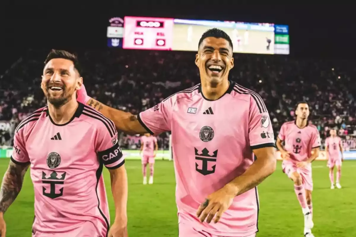 Two players smile and celebrate on the field wearing Inter Miami’s pink uniform during a nighttime match.