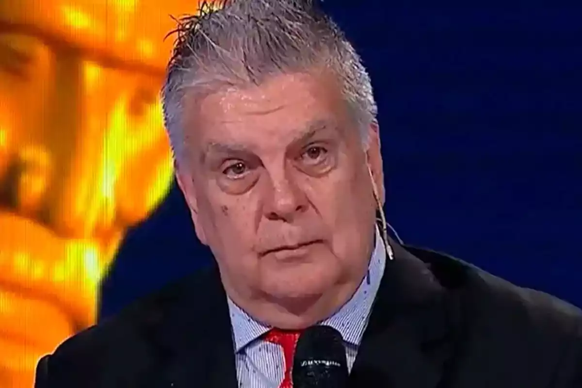 An older man with gray hair and a serious expression holds a microphone while on a lit stage. An older man with gray hair and a serious expression holds a microphone while on a lit stage.