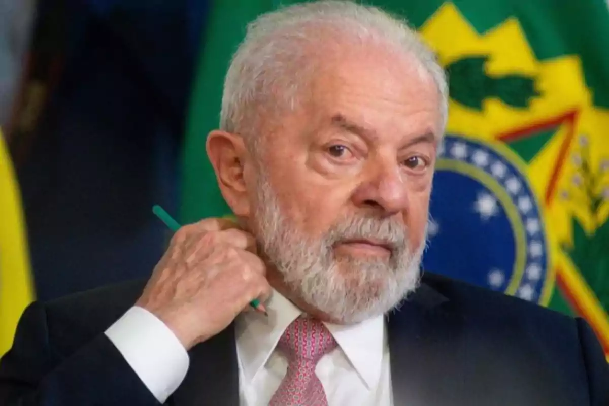 Un hombre mayor con barba blanca y traje oscuro sostiene un bolígrafo verde frente a una bandera de Brasil.