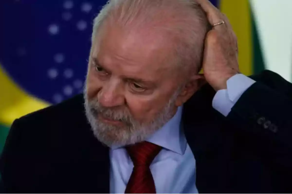 Un hombre mayor con barba y cabello canoso se toca la cabeza mientras viste traje y corbata roja con una bandera de Brasil de fondo.