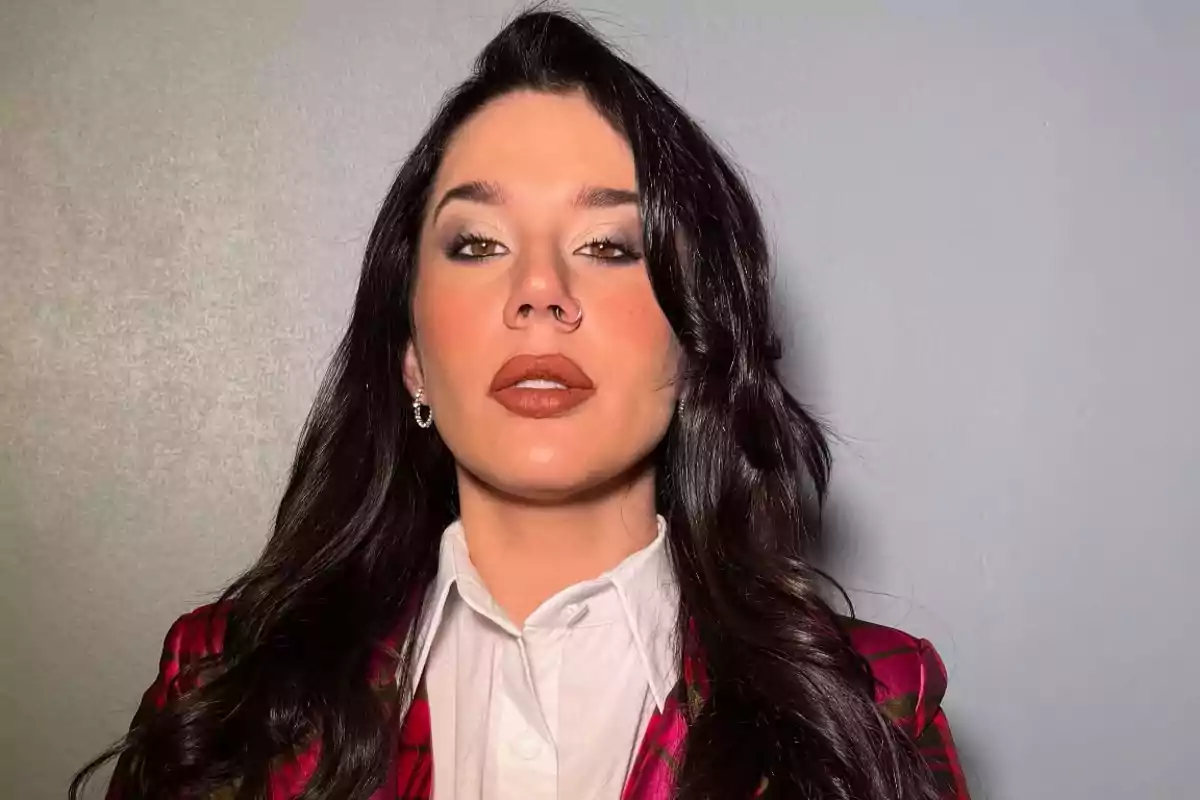 Mujer de cabello largo y oscuro con maquillaje llamativo, usando una camisa blanca y saco rojo, posando frente a una pared gris