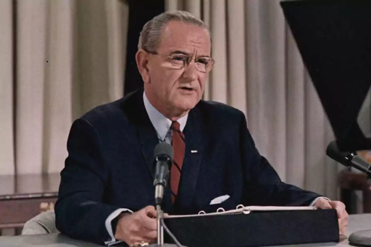 An older man with glasses and a dark suit is speaking in front of a microphone in a formal setting. An older man with glasses and a dark suit is speaking in front of a microphone in a formal setting.