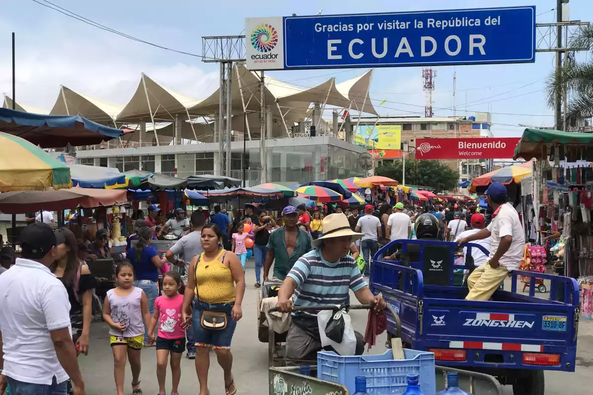 Multitud de personas caminando entre puestos de mercado al aire libre en una zona fronteriza entre Ecuador y Perú con letreros de bienvenida y despedida visibles sobre la calle