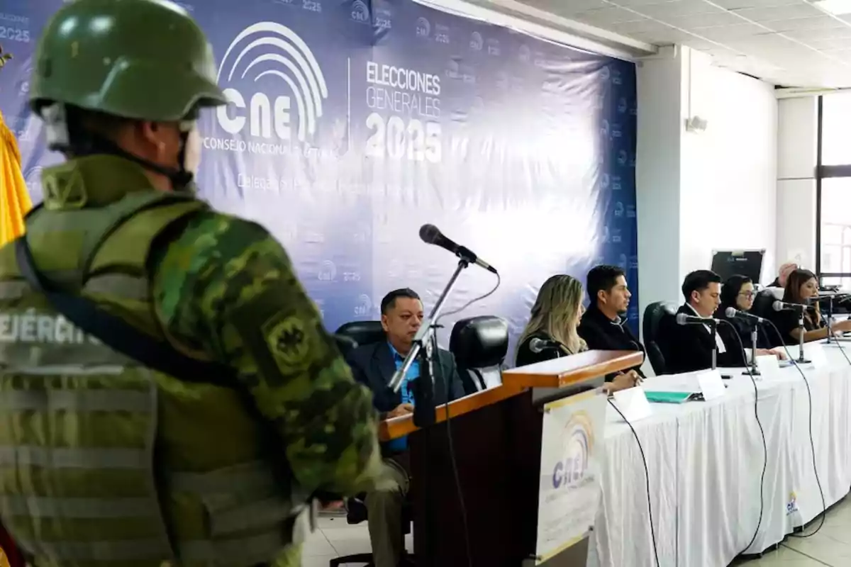 A soldier with his back turned observes a table with several people seated in front of microphones at a National Electoral Council event with a background that says "General Elections 2025."