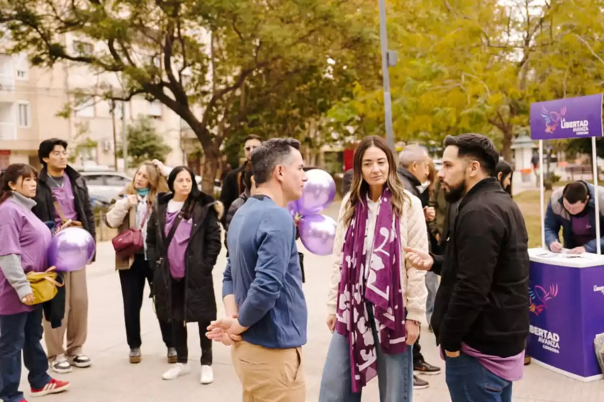 Personas reunidas al aire libre en una plaza, algunas con globos y prendas moradas, conversando cerca de un puesto informativo Personas reunidas al aire libre en una plaza, algunas con globos y prendas moradas, conversando cerca de un puesto informativo